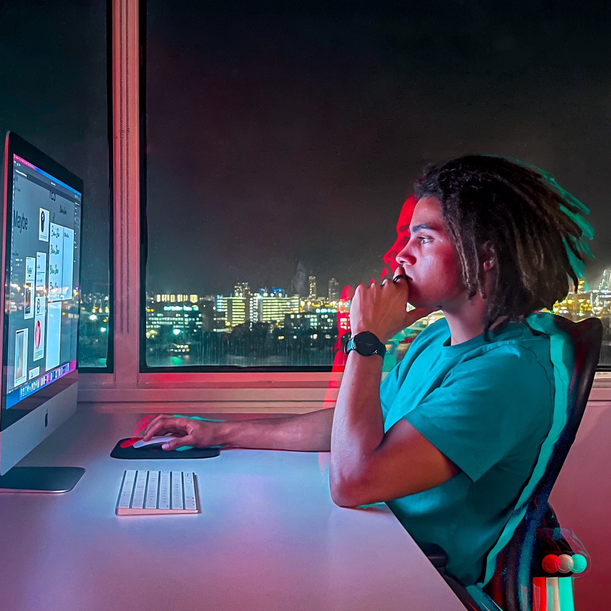 Talon Smith at his desk at night, looking at his iMac with a city skyline behind him